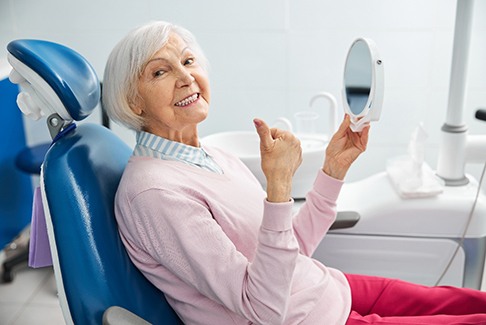 Senior woman sitting in dental chair, holding mirror, and giving thumbs up